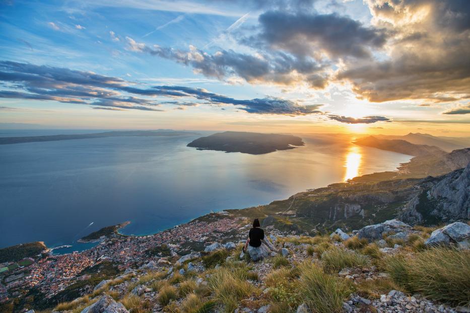 Eine Person sitzt auf einem Felsen und blickt auf die Küstenstadt und das Meer bei Sonnenuntergang.