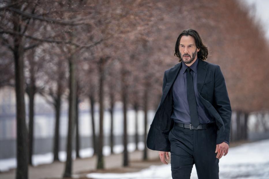 Keanu Reeves in einem dunklen Anzug geht auf einem verschneiten Weg entlang einer Baumreihe.