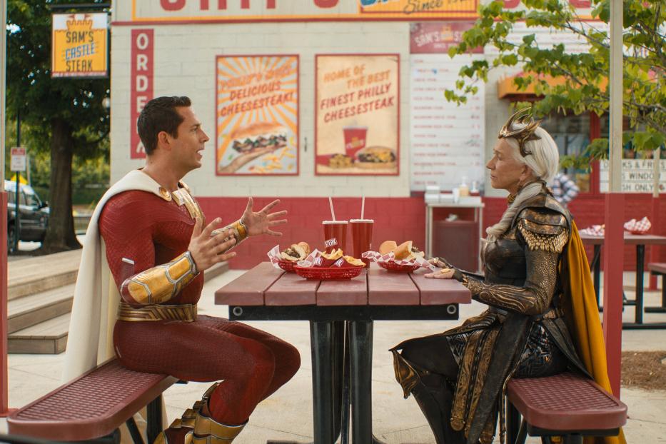 Shazam und Hespera sitzen an einem Tisch vor einem Imbiss und essen Cheesesteaks.
