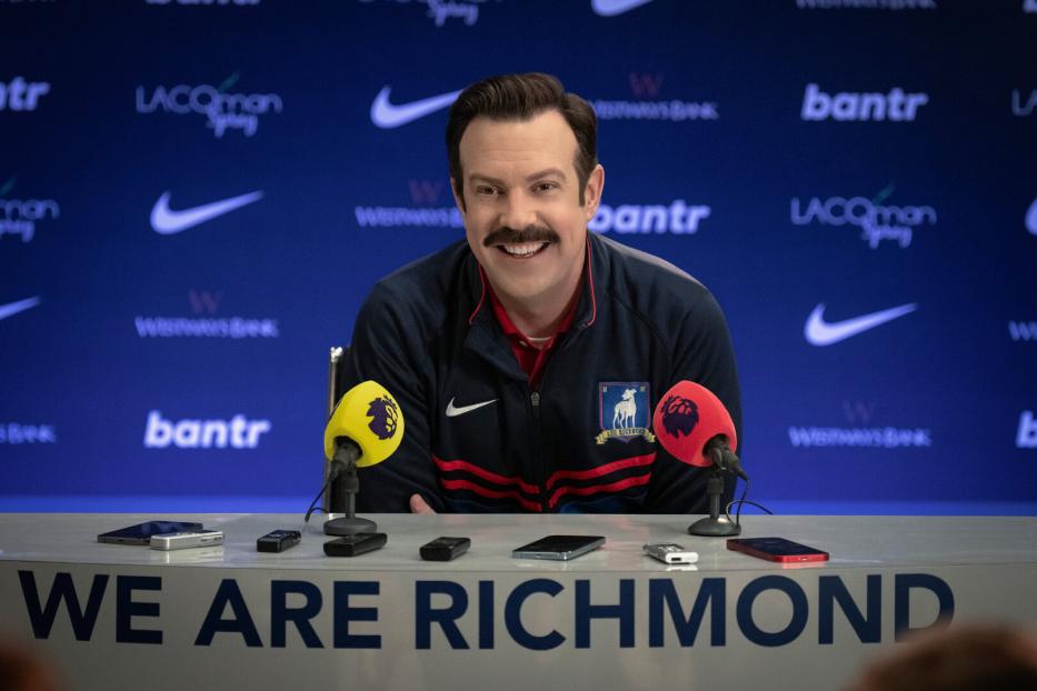 Ted Lasso sitzt lächelnd bei einer Pressekonferenz von AFC Richmond.