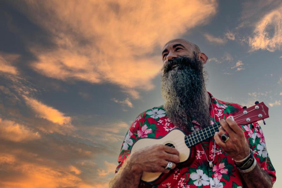 Rasierter Mann mit langem Bart spielt Ukulele vor einem dramatischen Himmel.