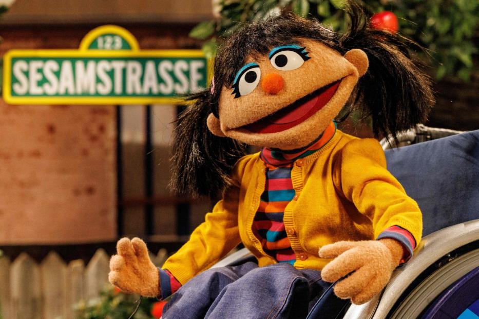 Eine Puppe der „Sesamstraße“ sitzt in einem Rollstuhl vor dem Straßenschild.