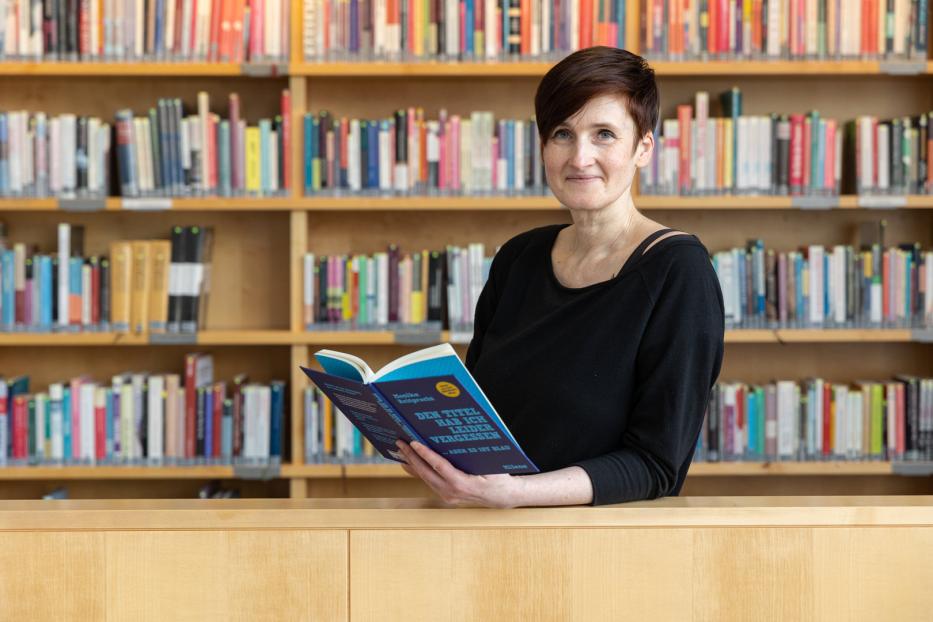 Eine Frau liest in einer Bibliothek das Buch „Den Titel hab ich leider vergessen… aber es ist blau“.