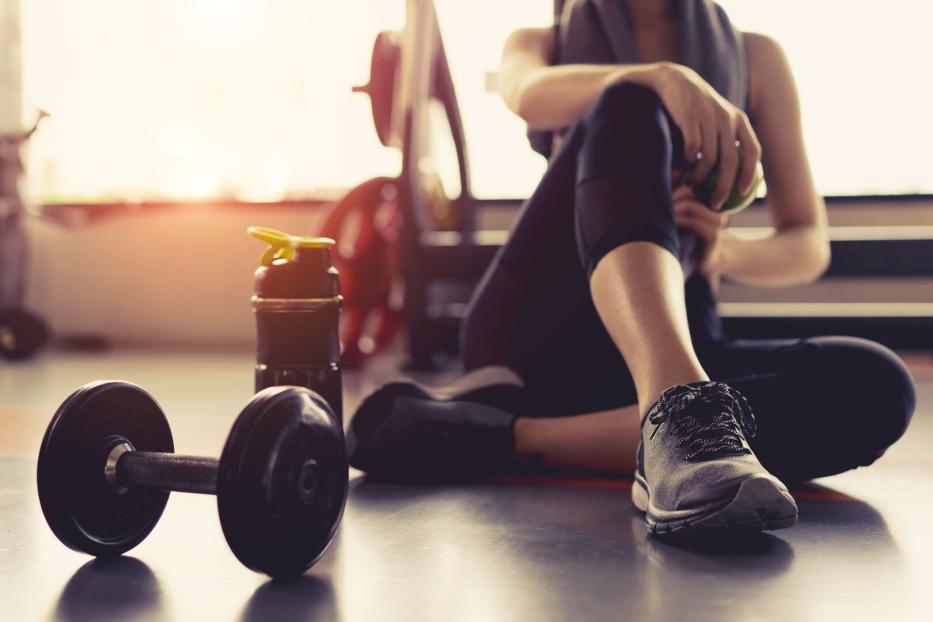 Eine Frau sitzt im Fitnessstudio mit einer Hantel und einer Wasserflasche neben sich.