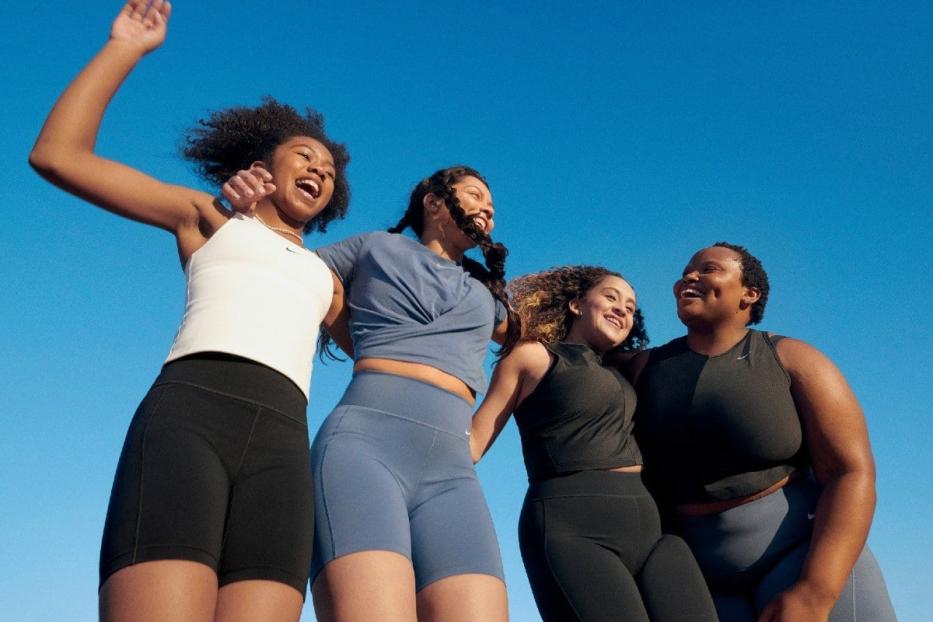 Vier Frauen in Sportkleidung von Nike stehen zusammen und lachen vor blauem Himmel.