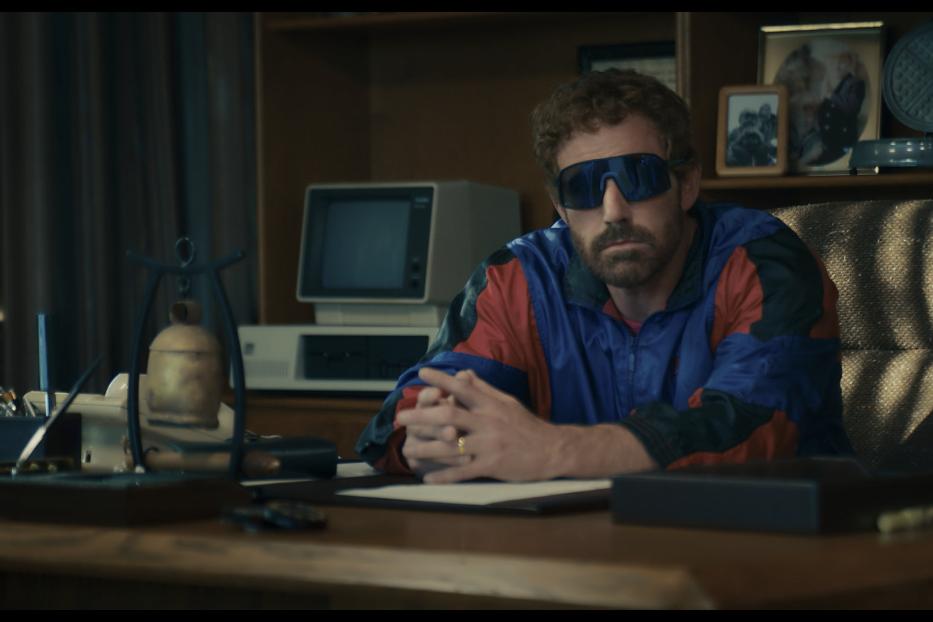 Ben Affleck sitzt mit Sonnenbrille an einem Schreibtisch mit einem alten Computer.