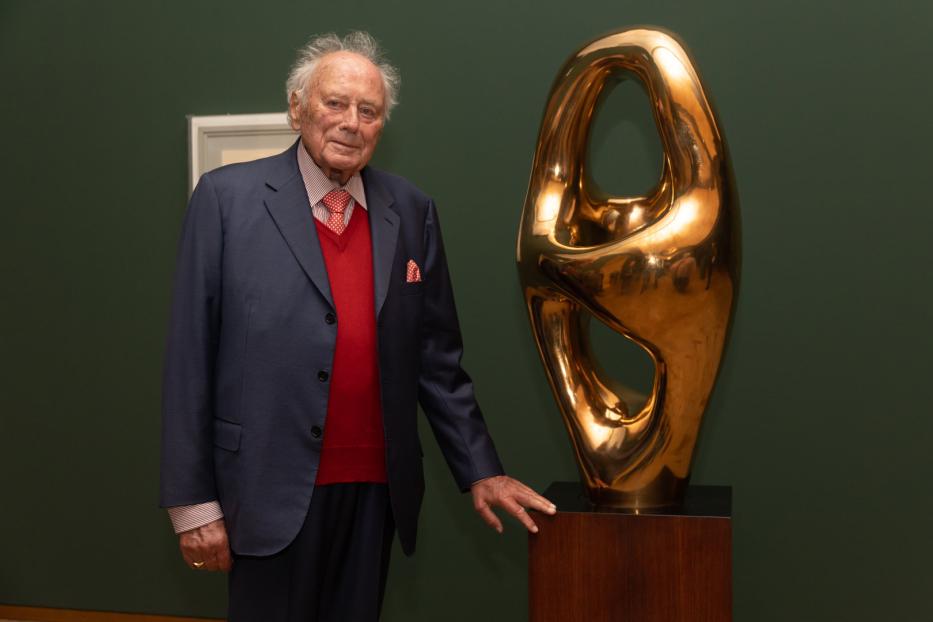 Ein älterer Mann posiert neben einer abstrakten, goldenen Skulptur.