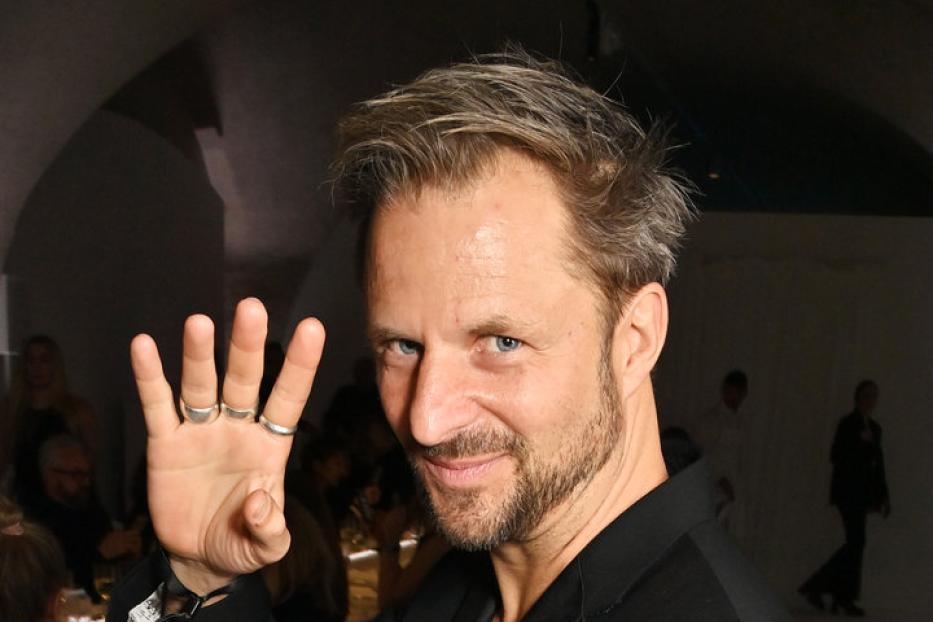 Ein Mann mit Anzug und mehreren Ringen zeigt mit der Hand eine Vier.