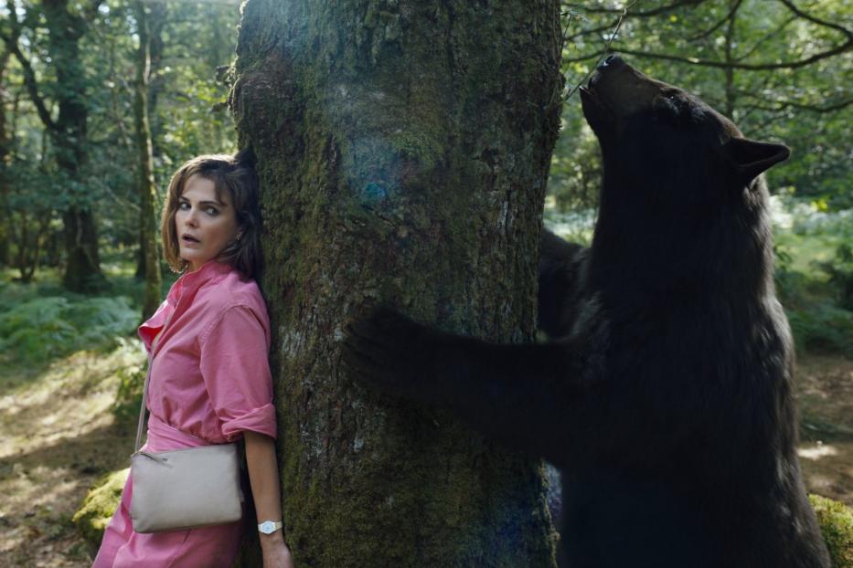 Eine Frau in einem Wald lehnt ängstlich an einem Baum, während sich ein Bär nähert.