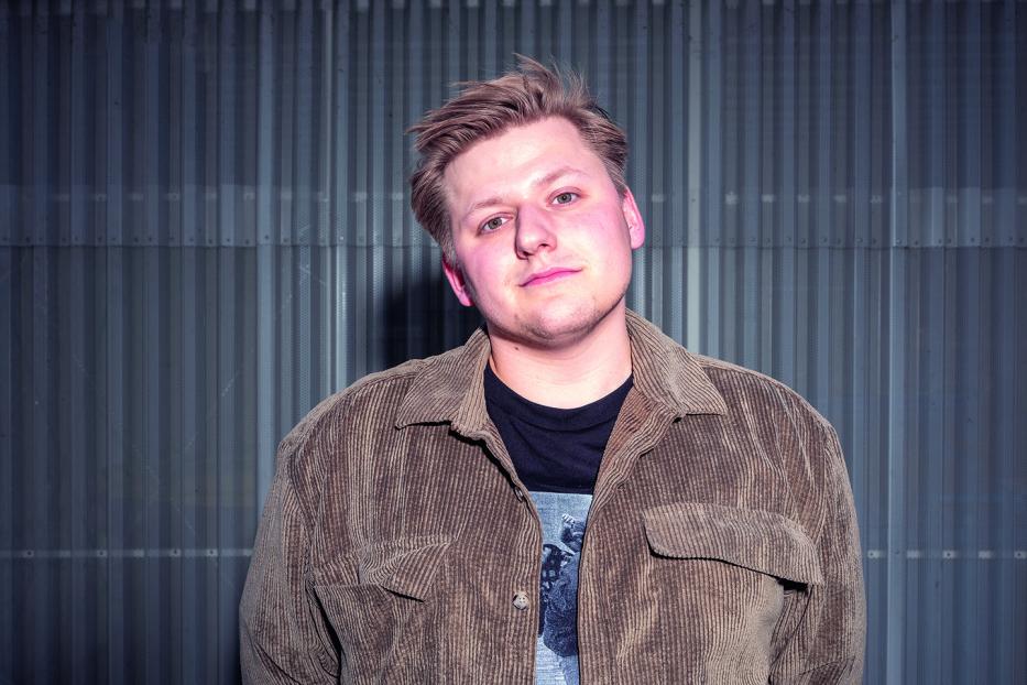 Ein junger Mann mit blonden Haaren und brauner Cordjacke vor einer grauen Wand.