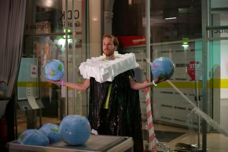 Ein Mann in einem Kostüm aus Müll und Styropor hält zwei Globen in den Händen.