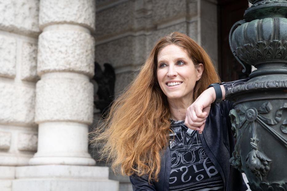 Eine Frau mit langen, roten Haaren lehnt an einer verzierten Laterne vor einem Gebäude.
