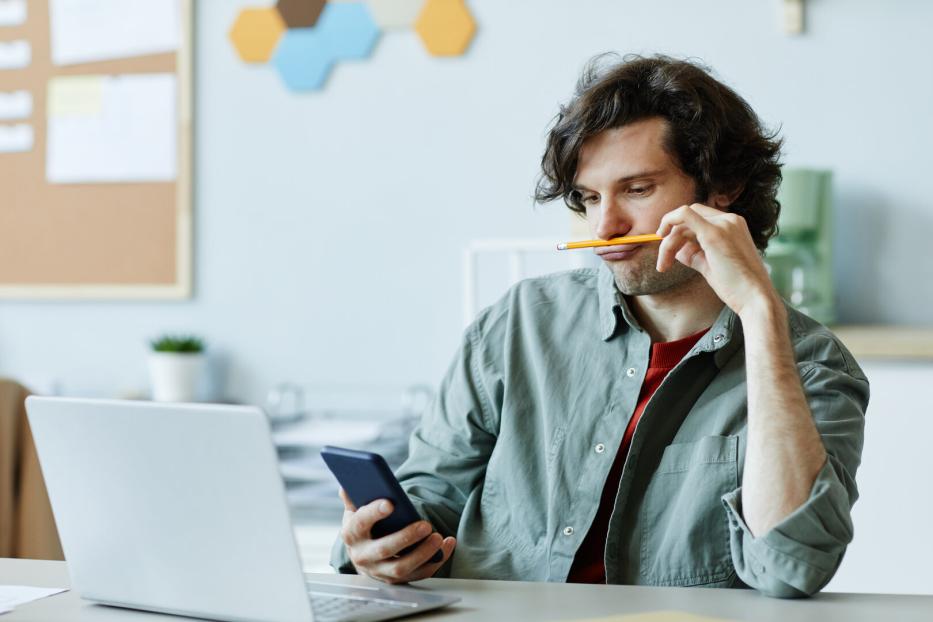 Ein Mann sitzt mit einem Stift im Mund vor einem Laptop und schaut auf sein Handy.