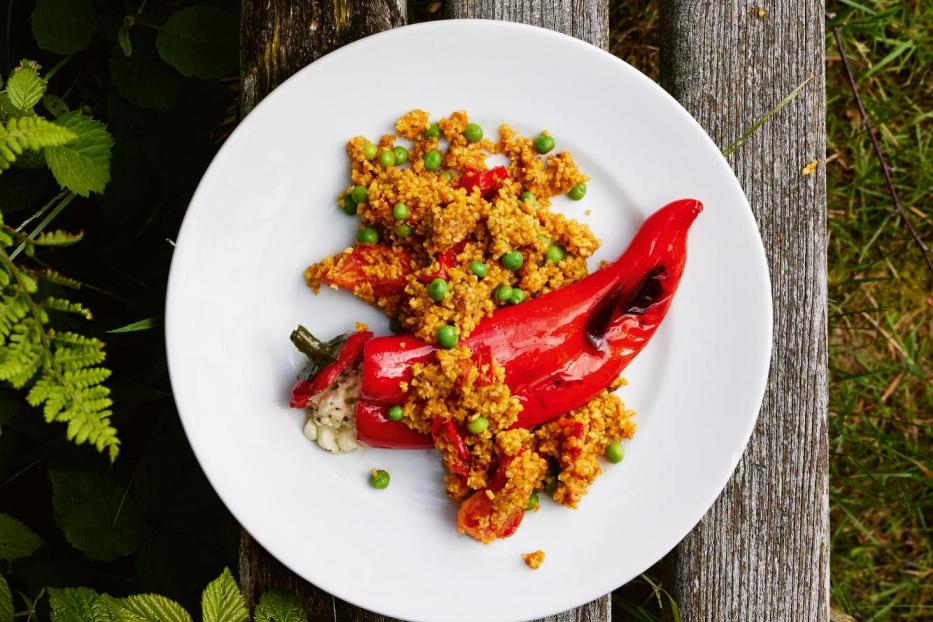Ein Teller mit gefüllter Paprika und Couscous steht auf einer Holzbank neben einem Bier.