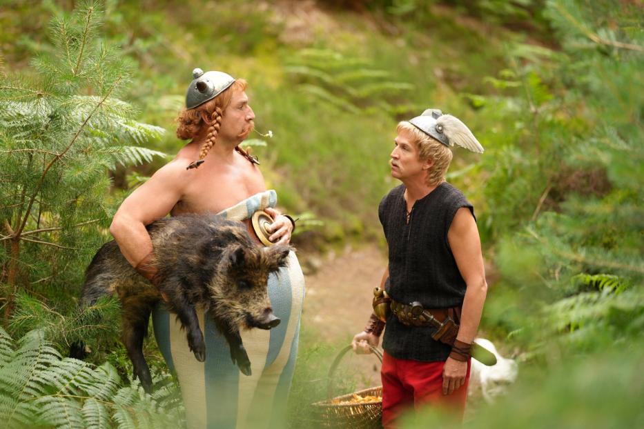 Asterix trägt ein Wildschwein, während Obelix mit einem Korb Pilze daneben steht.