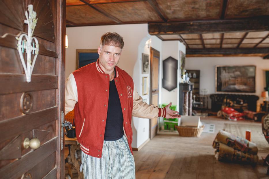 Ein Mann in Collegejacke und Pyjamahose steht in einer offenen Tür.