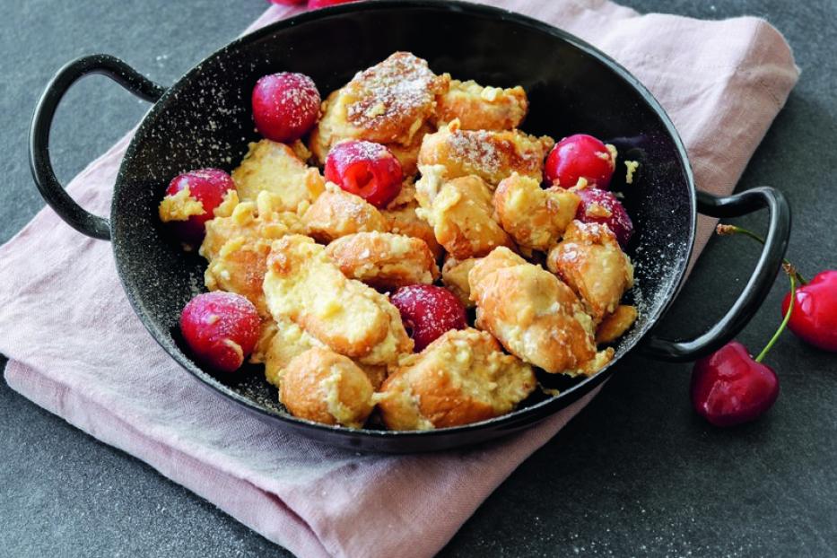 Kaiserschmarrn mit Kirschen in einer schwarzen Pfanne auf einem rosa Tuch angerichtet.