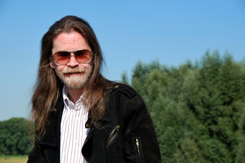 Ein Mann mit langen Haaren, Bart und Sonnenbrille vor blauem Himmel und Bäumen.