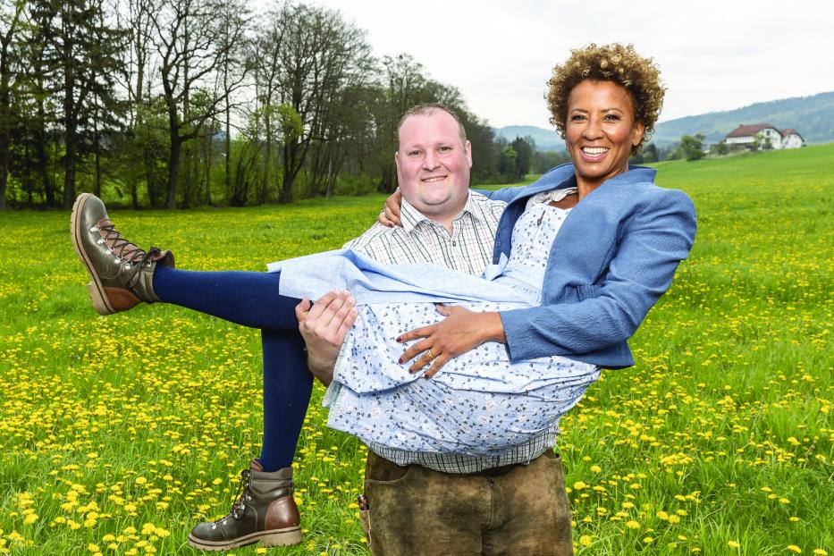 Arabella Kiesbauer wird von einem Mann auf einer Wiese getragen.