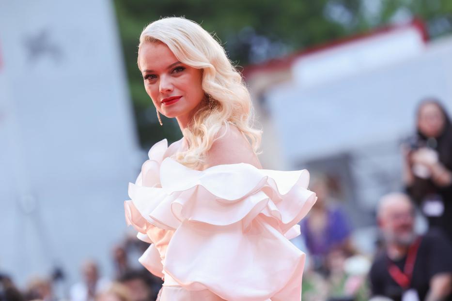 Eine Frau mit blonden, gewellten Haaren und einem rosa Kleid mit Rüschen.
