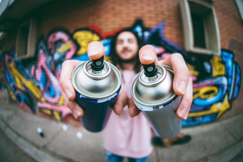 Eine Person hält zwei Sprühdosen vor einer mit Graffiti bemalten Wand.