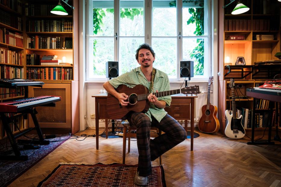 Ein Mann mit Gitarre sitzt in einem Raum voller Bücher und Musikinstrumente.