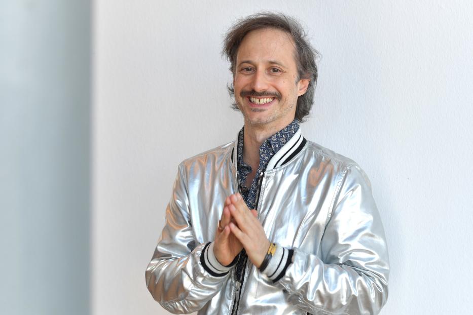 Ein lächelnder Mann mit Schnurrbart und silberner Jacke steht vor einer weißen Wand.