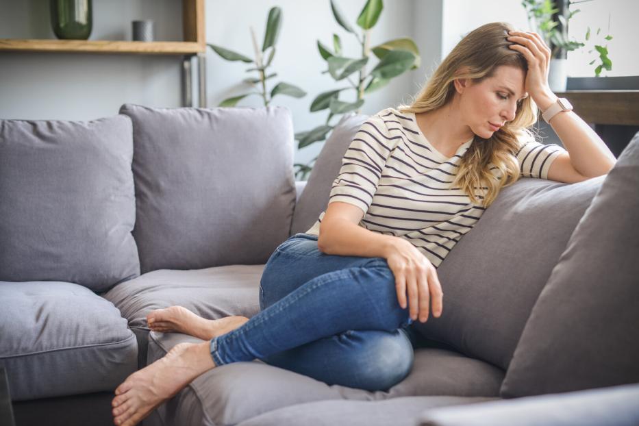 Eine Frau mit blonden Haaren sitzt nachdenklich auf einem grauen Sofa.
