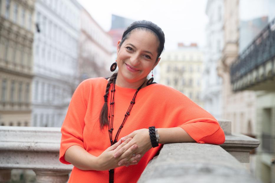 Eine lächelnde Frau in einem orangefarbenen Pullover lehnt an einer Balustrade.