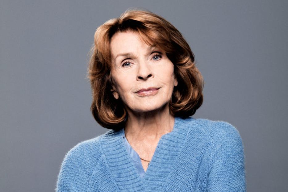 Eine ältere Frau mit verschränkten Armen trägt eine blaue Strickjacke.