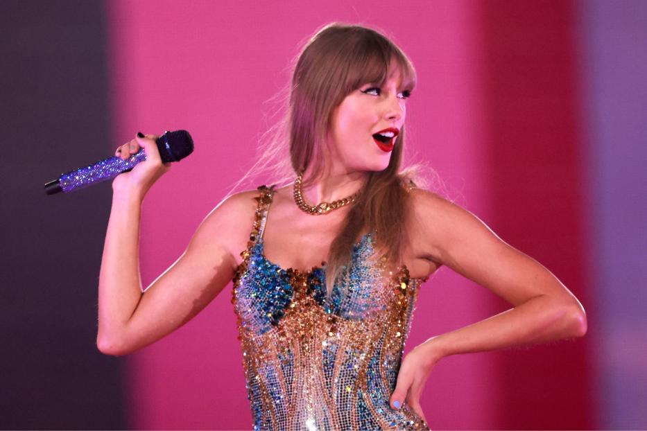 Taylor Swift auf der Bühne mit einem Mikro in der Hand, schaut verschmitzt.