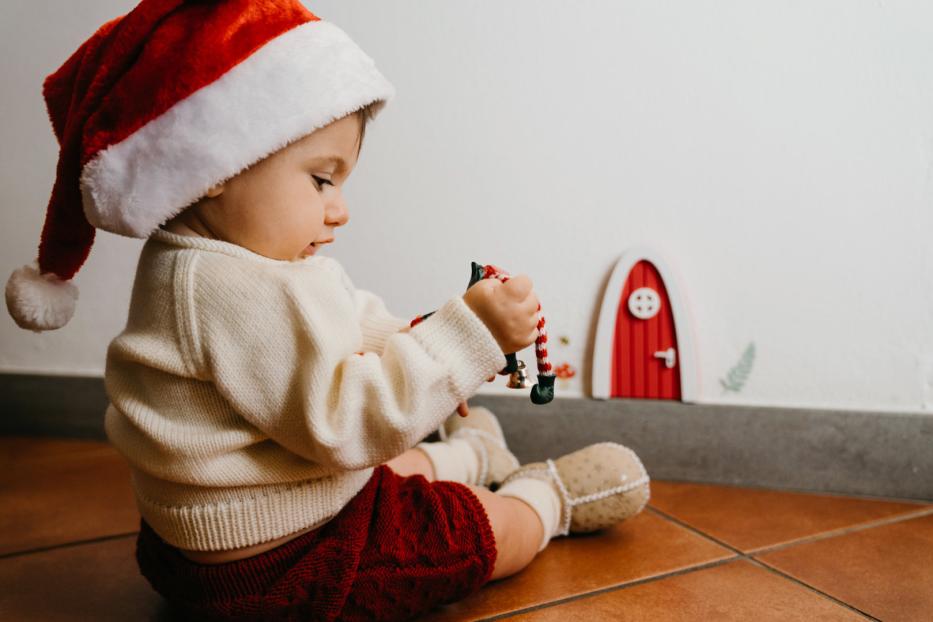 Ein Baby mit Weihnachtsmütze spielt mit einer kleinen Elfenpuppe vor einer roten Tür.