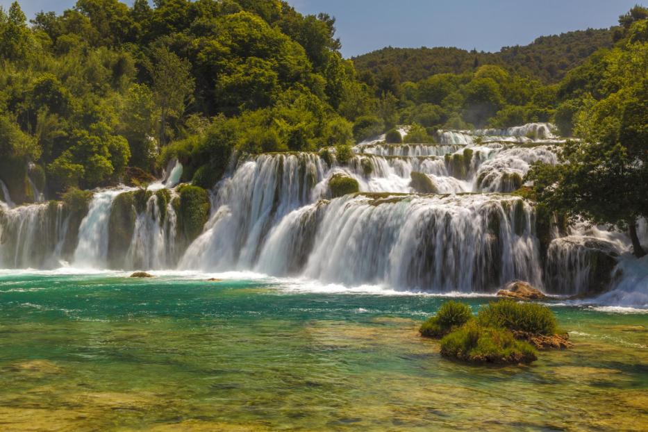Mehrere Wasserfälle ergießen sich in einen türkisfarbenen See, umgeben von grünen Bäumen.