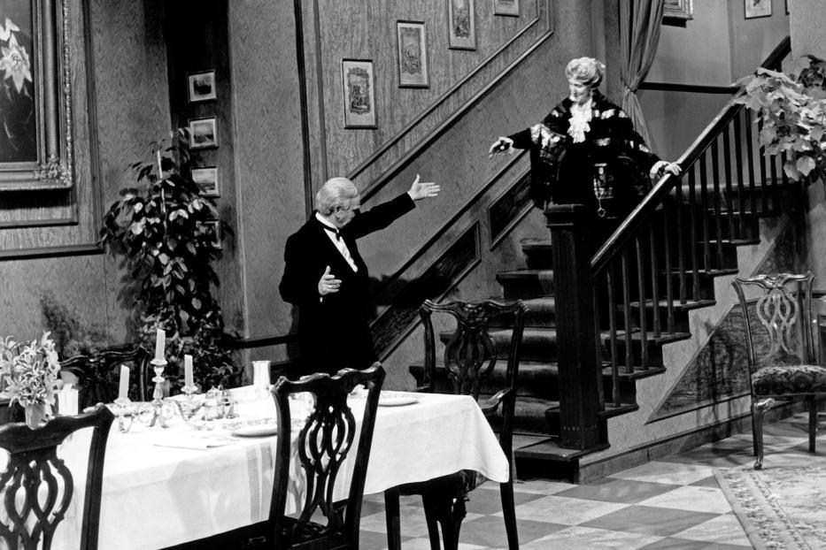 Ein Mann im Smoking begrüßt eine Frau auf einer Treppe in einem eleganten Haus.