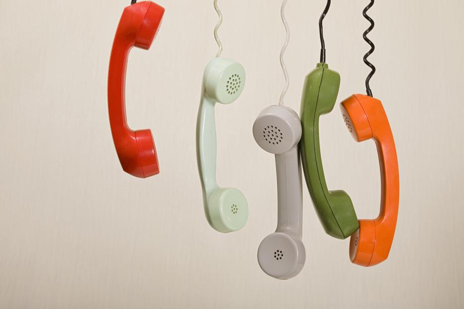 Mehrere farbige Telefonhörer hängen an ihren Kabeln vor einem beigen Hintergrund.