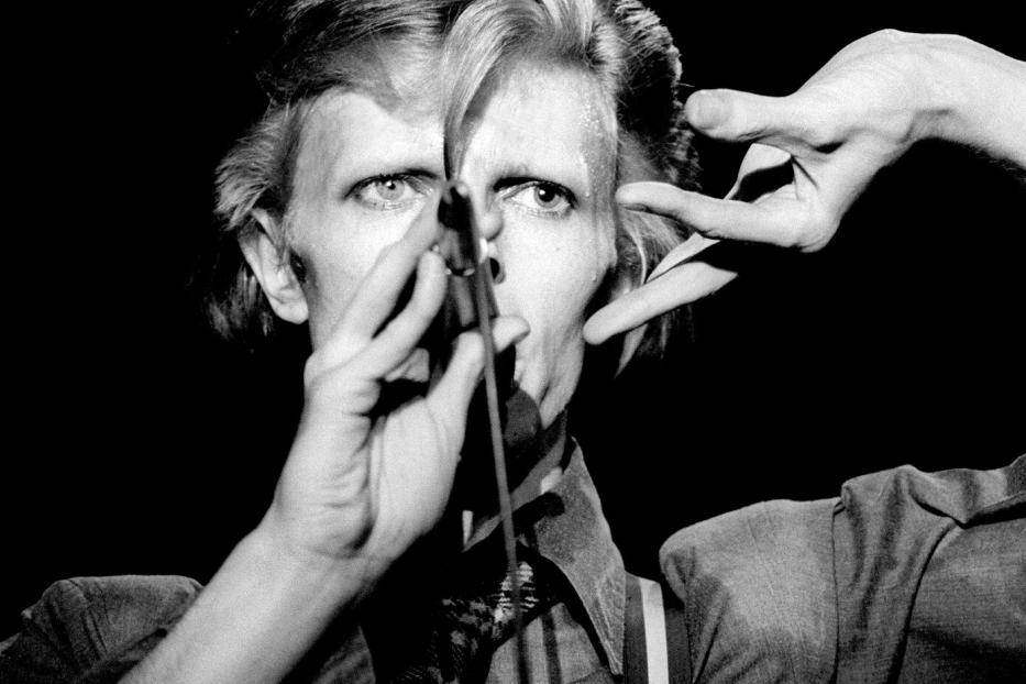 David Bowie singt in einem dunklen Raum mit einem Mikrofon vor dem Gesicht.