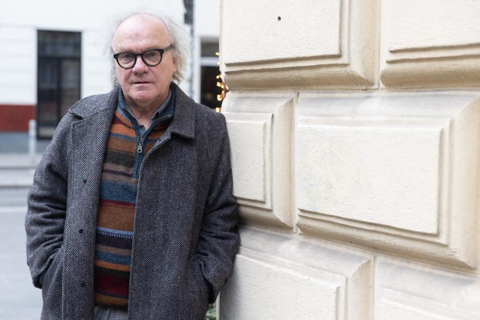 Ein Mann mit Brille und grauem Haar lehnt an einer hellen Wand.