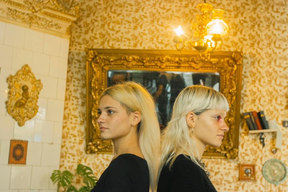 Zwei junge Frauen mit blonden Haaren stehen Rücken an Rücken vor einer goldenen Wand.