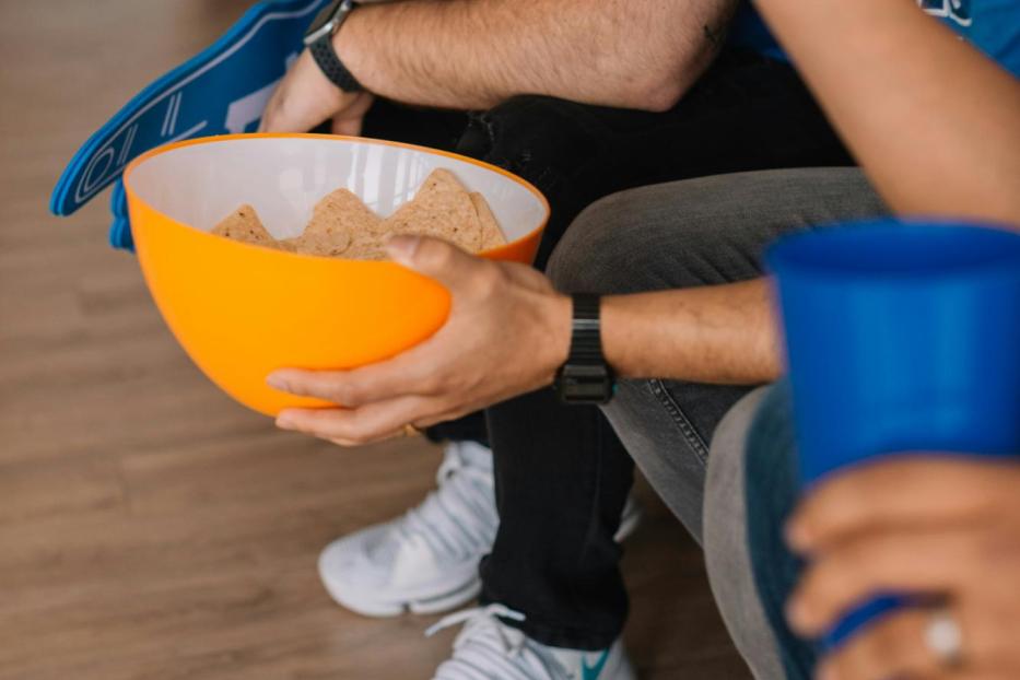 Zwei Männer in blauen T-Shirts schauen ein Spiel und essen Tortilla-Chips.