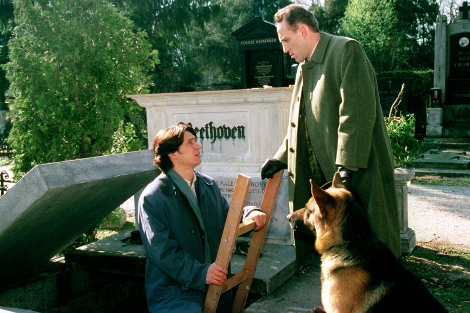 Auf einem Friedhof stehen ein Mann mit Leiter, ein anderer Mann und ein Schäferhund vor einem Grabstein mit der Aufschrift „Beethoven“.