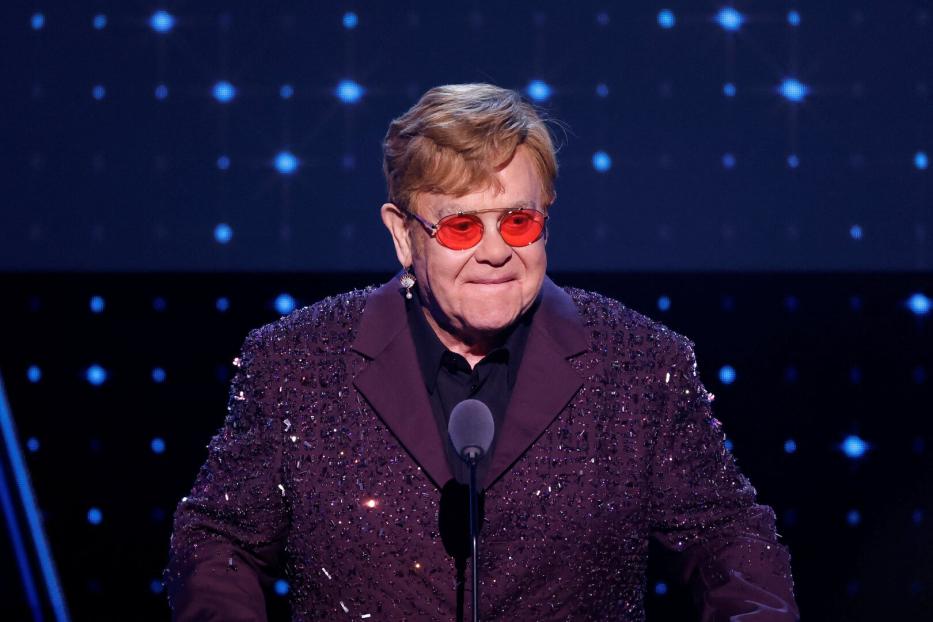 Elton John steht an einem Mikrofon und trägt eine rote Brille.