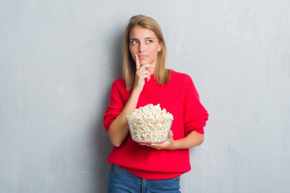 Eine Frau im roten Pullover hält eine Schüssel Popcorn und schaut nachdenklich zur Seite.
