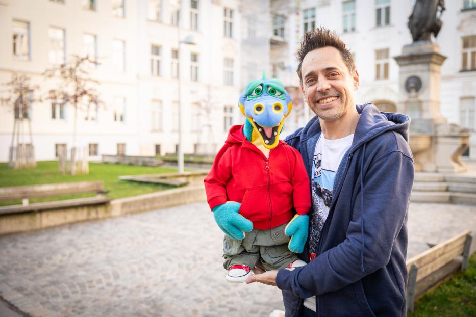 Ein Mann lächelt mit einer bunten Drachenpuppe im Arm vor einem Gebäude.