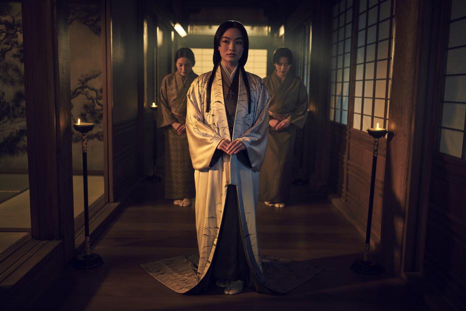 Eine Frau in einem Kimono steht in einem traditionellen japanischen Haus, flankiert von zwei weiteren Frauen.