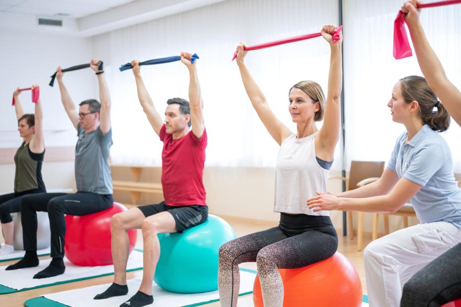 Eine Gruppe von Menschen sitzt auf Gymnastikbällen und dehnt sich mit Bändern unter Anleitung einer Physiotherapeutin.