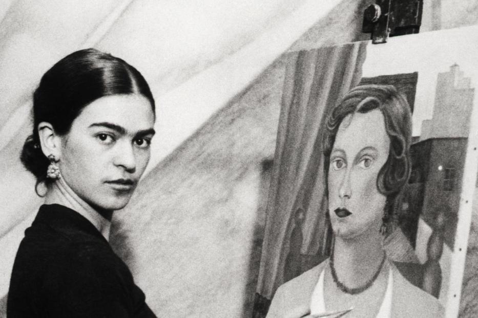 Frida Kahlo steht vor einer Staffelei und malt ein Porträt.