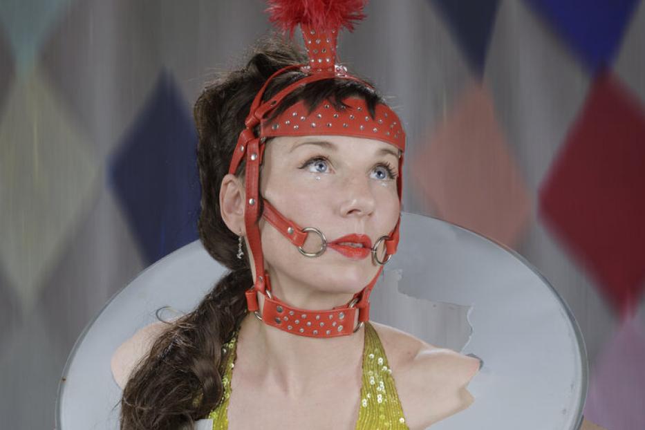 Eine Frau mit einem roten Federkopfschmuck und einem ungewöhnlichen Kragen vor einem bunten Hintergrund.