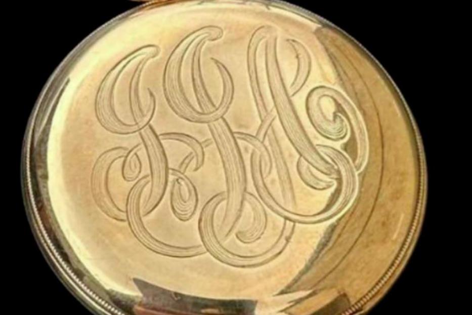Eine goldene Taschenuhr mit eingraviertem Monogramm vor schwarzem Hintergrund.