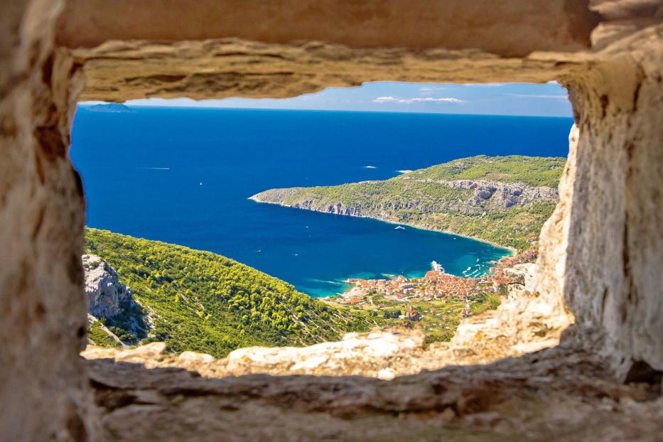Blick durch eine steinerne Fensteröffnung auf eine Küstenstadt und das Meer.