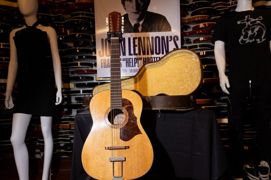 John Lennons Gitarre steht vor einem Poster von ihm und einem Gitarrenkoffer.